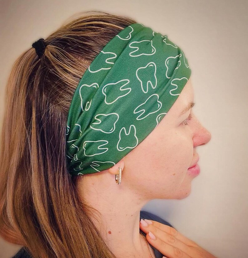 May include: A wide fabric headband in a deep forest green color featuring a repeating white line art pattern of human teeth. The headband is worn across the forehead and is made of a smooth, flexible material that fits snugly around the head.
