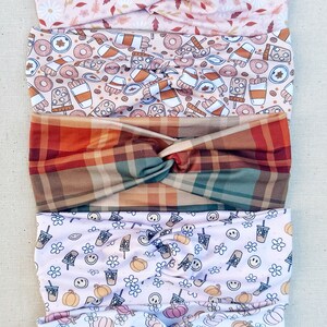 May include: Five different patterned headbands stacked on top of each other. The top headband is pink with white flowers and brown leaves. The second headband is beige with brown coffee cups, donuts, and cinnamon sticks. The third headband is a plaid pattern with red, brown, and green squares. The fourth headband is pink with white flowers, smiley faces, and brown coffee cups. The bottom headband is white with pink and orange pumpkins.