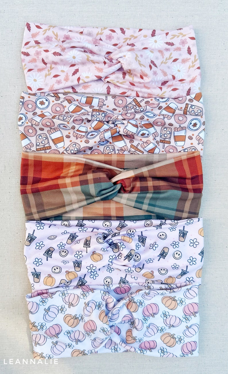 May include: Five different patterned headbands stacked on top of each other. The top headband is pink with white flowers and brown leaves. The second headband is beige with brown coffee cups, donuts, and cinnamon sticks. The third headband is a plaid pattern with red, brown, and green squares. The fourth headband is pink with white flowers, smiley faces, and brown coffee cups. The bottom headband is white with pink and orange pumpkins.