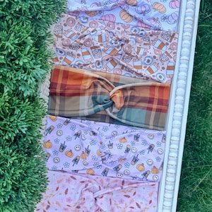 May include: Five fabric headbands with twisted knots, featuring various patterns including purple and orange pumpkins, coffee cups and donuts, brown and orange plaid, light purple with pumpkins and smiley faces, and pink floral autumn leaves.