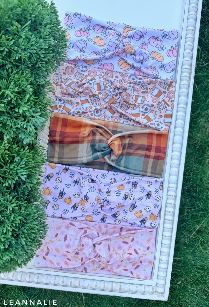 May include: Five fabric headbands with twisted knots, featuring various patterns including purple and orange pumpkins, coffee cups and donuts, brown and orange plaid, light purple with pumpkins and smiley faces, and pink floral autumn leaves.