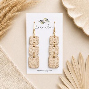 May include: Gold-toned dangle earrings with three square, beige leopard print beads. The earrings hang from gold-colored hooks and are displayed on a white card with the text "Leannalie" and "Leannalie.Etsy.com".
