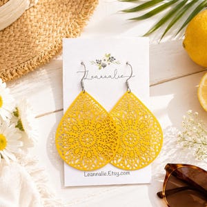 May include: A pair of bright yellow, teardrop-shaped earrings with intricate cut-out designs. The earrings hang from silver hooks and are displayed on a white card with the text "Leannalie.Etsy.com". The earrings are next to a straw hat, daisies, and sunglasses.