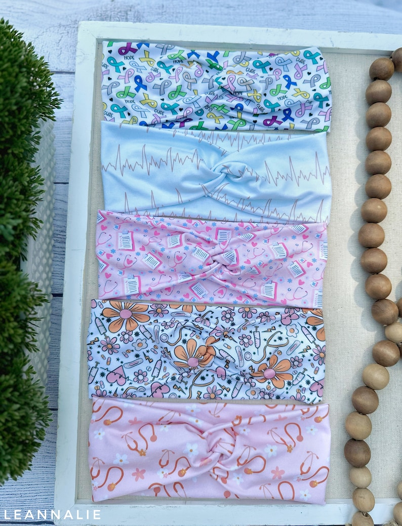 K&ouml;nnte beinhalten: Vier verschiedene gemusterte Stirnb&auml;nder mit einem Knotendesign. Die Stirnb&auml;nder werden auf einem wei&szlig;en Hintergrund mit einer Holzperlenkette pr&auml;sentiert. Die Muster umfassen rosa und wei&szlig; mit einem Stethoskop-Design, wei&szlig; mit einem floralen und medizinischen Design, rosa mit einem Stethoskop-Design und wei&szlig; mit einem Band-Design.