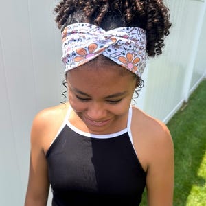 May include: A white fabric headband featuring a twisted front design and a repeating pattern of orange flowers, medical syringes, and small black symbols. The headband is worn across the forehead, contrasting with the black sleeveless top worn below.