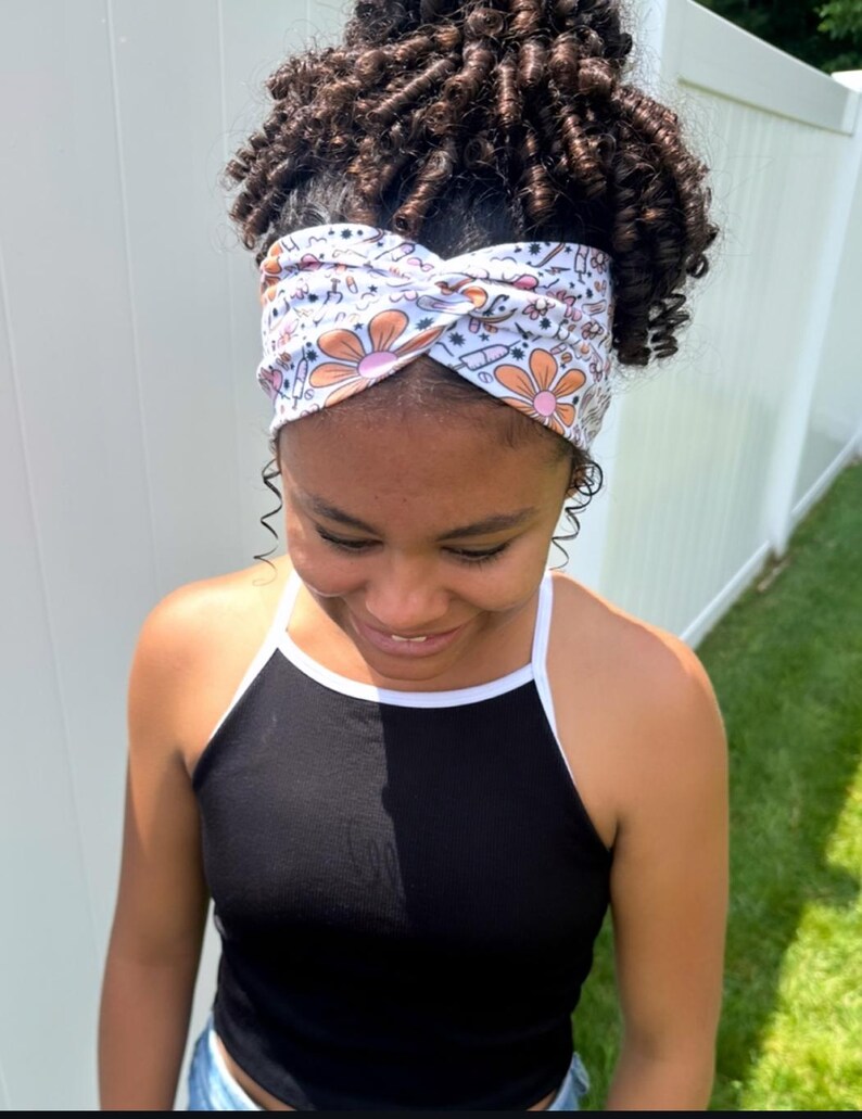 May include: A white fabric headband featuring a twisted front design and a repeating pattern of orange flowers, medical syringes, and small black symbols. The headband is worn across the forehead, contrasting with the black sleeveless top worn below.