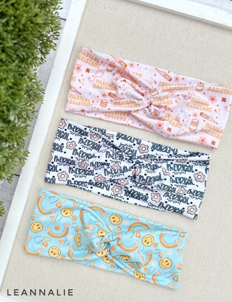 May include: Three colorful headbands with different patterns. The first headband is pink with a white pattern of medical symbols and the text "Medical Assistant". The second headband is white with a black pattern of flowers and the text "Medical Assistant". The third headband is light blue with a yellow pattern of rainbows and smiley faces.