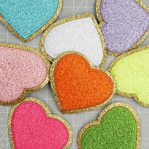 Op de afbeelding: Een verzameling hartvormige patches in verschillende kleuren met een getextureerde, pluche uitstraling. Elk hart is omrand met gouden glitter. Kleuren zijn blauw, wit, roze, oranje, paars, geel, groen en felroze.