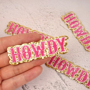 May include: Four pink and gold glitter iron-on patches with the word "HOWDY" in a stylized font.