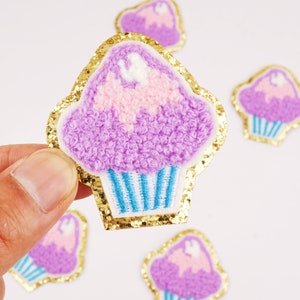 Op de afbeelding: Geborduurde cupcake-patches met een gouden glitterrand. De cupcakes hebben paarse glazuur, roze en witte accenten en blauw-wit gestreepte cupcake-vormpjes. Ideaal voor het versieren van kleding of accessoires.