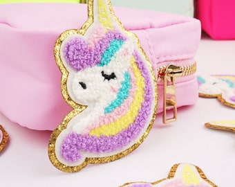 May include: A pink zippered pouch with a gold zipper and a fluffy purple, white, and yellow unicorn patch. The unicorn patch has a gold glitter outline.
