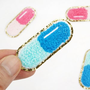 May include: A blue and light blue pill shaped iron-on patch with gold glitter trim. The patch is made of fuzzy fabric.