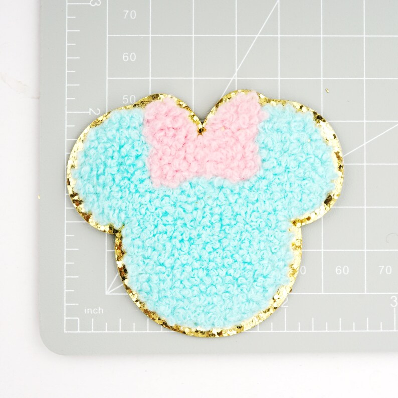 Peut inclure: Une applique en tissu pelucheux bleu clair et rose avec un contour de paillettes dor&eacute;es en forme de t&ecirc;te de souris de dessin anim&eacute;.