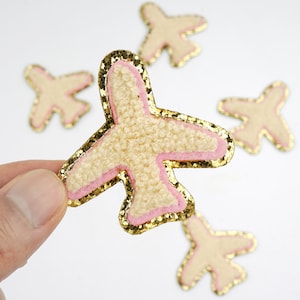 Puede incluir: Un conjunto de parches con forma de avión. Los parches presentan un centro texturizado de color beige claro, delineado con rosa y purpurina dorada. Un parche es sostenido por una mano, con otros dispersos en el fondo.