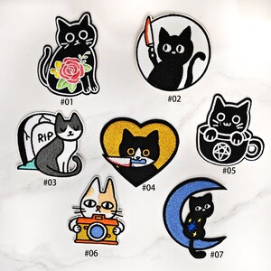 May include: Set of seven iron-on embroidered patches featuring black cats in various poses. The patches include a cat with a rose, a cat holding a knife, a cat with a tombstone, a cat holding a knife in a heart, a cat in a teacup, a cat with a camera, and a cat on a crescent moon.
