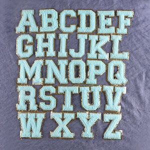 May include: A set of 26 light blue felt letters with gold trim, arranged in alphabetical order. The letters are uppercase and have a classic varsity style.