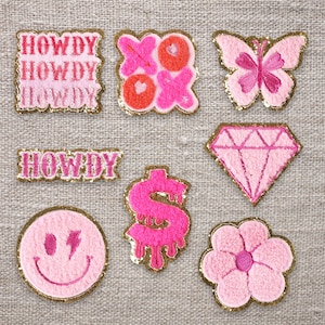 Puede incluir: Un conjunto de diez parches termoadhesivos de color rosa y dorado con varios diseños, que incluyen un diamante, una flor, una mariposa, un signo de dólar, una cara sonriente y la palabra "Howdy" en diferentes estilos.