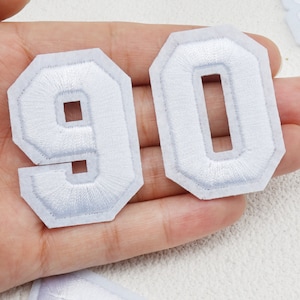 Può includere: Due toppe ricamate bianche, una con il numero 9 e l'altra con il numero 0, che formano il numero 90. I numeri sono in un classico font collegiale, con un bordo bianco.