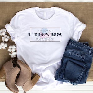 May include: White t-shirt with the text "If she smokes cigars she's a real one" printed on it. The shirt is laying on a brown surface with a pair of brown boots and blue jeans.