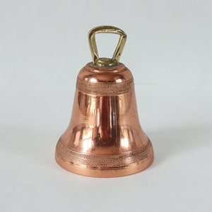 Op de afbeelding: Een kleine koperen bel met een messing handvat. De bel heeft een decoratief patroon rond de rand.