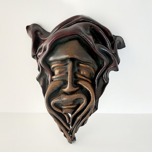 May include: A brown leather wall hanging depicting a stylized face with a beard and hooded cloak. The face has a prominent nose and a detailed, textured surface. The color palette includes shades of brown and reddish-brown.