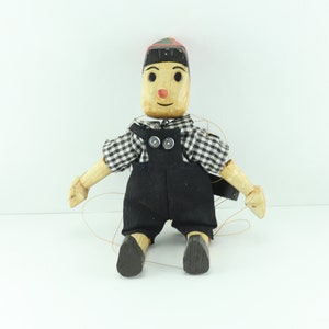 May include: A wooden puppet with a black and white checkered shirt, black overalls, and a red and green hat. The puppet has a painted face with a red nose and black eyes.
