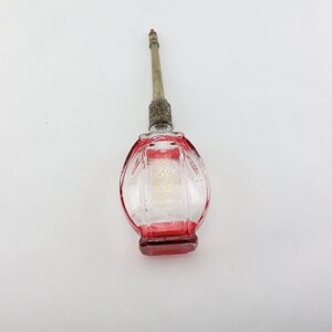 Puede incluir: Un frasco de perfume de vidrio transparente con base y detalles en rojo. El frasco tiene forma ovalada con estrías verticales y una tapa y aplicador decorativos de metal plateado.