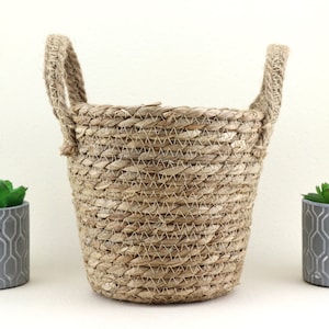 Op de afbeelding: Een natuurlijke geweven zeegrasmand met twee touwhandvatten. De mand is lichtbruin en heeft een ronde vorm.