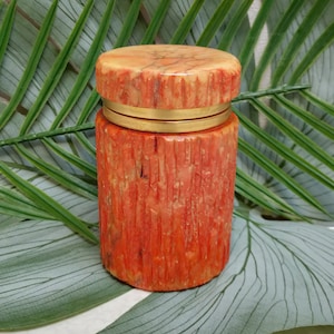 May include: A cylindrical, coral-colored container with a matching lid. The container has a textured, vertical-lined pattern. A gold-colored band encircles the container near the top. The lid is a lighter shade of coral with a marbled appearance. The container is likely used for storage.