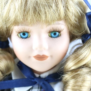 Puede incluir: Una muñeca de porcelana con el pelo rubio peinado en trenzas con cintas azules. La muñeca tiene ojos azules y una sonrisa pintada.