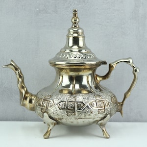 May include: Ornate silver teapot with embossed detailing. The teapot features a curved spout, a looped handle, and rests on three small feet. The design includes floral and geometric patterns. The teapot's lid is topped with a decorative finial, adding to its elegant appearance.