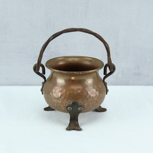 Puede incluir: Una pequeña olla de cobre con acabado martillado y tres patas de hierro. La olla tiene un asa de hierro retorcido.