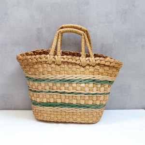 Op de afbeelding: Een geweven strooien tas met groene strepen. De tas heeft twee handvatten en is gemaakt van natuurlijke materialen.