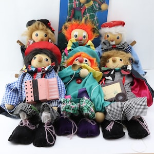 May include: A collection of handmade wooden marionette puppets. The puppets have painted faces, various hairstyles, and are dressed in colorful outfits. One puppet holds a pink accordion. Other puppets wear hats and bow ties.