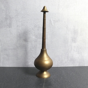 Puede incluir: Un frasco atomizador de metal dorado, alto y de base bulbosa, con un cuello largo y delgado. El frasco tiene una pequeña parte superior puntiaguda.