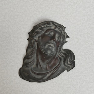 May include: A bronze-coloured relief sculpture of Jesus' face, wearing a crown of thorns. The sculpture shows long, flowing hair and a beard. The artist's signature, "M. Thomas," is in the lower left. The artwork is set against a light background.
