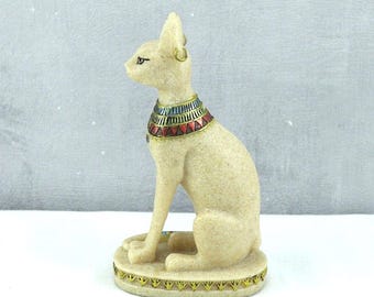 Puede incluir: Una figurita de cerámica blanca de un gato sentado con un collar dorado y azul. El gato tiene un pendiente dorado y está sentado sobre una base con un diseño dorado y blanco.