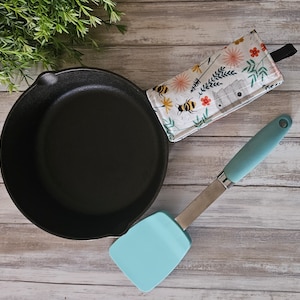 May include: A black cast iron skillet with a blue silicone spatula and a floral patterned pot holder with bees and flowers.
