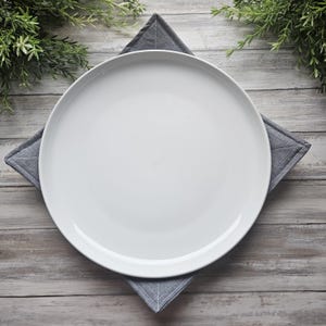 May include: A white ceramic plate rests on a gray, square quilted coaster. The plate is centered, with the coaster's corners visible. Greenery is arranged around the plate on a light wood-grain surface, creating a simple, elegant table setting.
