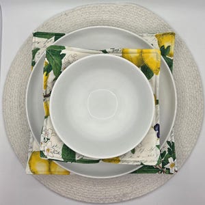 May include: A white ceramic bowl and plate set atop a lemon-themed placemat. The placemat features a pattern of yellow lemons, green leaves, and white flowers. The set rests on a round, woven, light gray placemat.