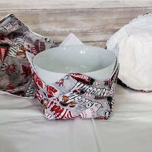 May include: Three white fabric bowl cozies with red and gray Wisconsin Badgers themed print. The cozies are folded and one is holding a white ceramic bowl.