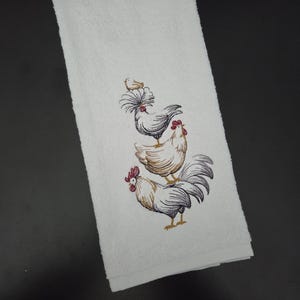 May include: White hand towel with an embroidered design of three roosters stacked on top of each other. The roosters are in shades of brown, black, and red. The towel is rectangular and made of a soft, absorbent material.