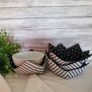 May include: Set of four black and white striped fabric bowl cozies. The cozies are folded and stacked, with one cozy holding a white ceramic bowl. The cozies have a black and white patterned fabric on the outside and a black fabric with white crosses on the inside.