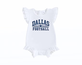 Pode incluir: Um body branco sem mangas com folhos nos ombros e nas aberturas das pernas. A frente apresenta o texto DALLAS FOOTBALL numa fonte azul desgastada com uma estrela azul e branca. Uma etiqueta no decote indica o tamanho 3-6M.