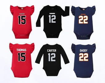 Può includere: Sei body per neonati con motivi sportivi. La fila superiore mostra body rossi, neri e blu navy con i numeri 15, 12 e 22. La fila inferiore mostra gli stessi colori con i nomi Thomas, Carter e Daddy sopra i numeri.