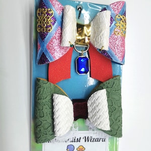 Könnte beinhalten: Zwei dekorative Haarschleifen. Die obere Schleife hat rosa Glitzer, rote, weiße und blaue Akzente sowie einen blauen Schmuckanhänger. Die untere Schleife hat grüne und weiße strukturierte Details. Die Schleifen sind auf einer blauen Rückseite befestigt.