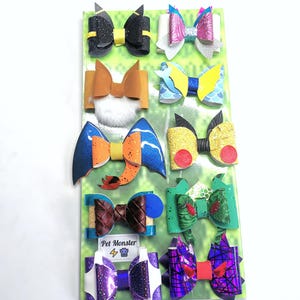 Może przedstawiać: Kolekcja kolorowych kokardek do włosów dla zwierząt domowych w różnych wzorach, w tym brokat, wzory i tekstury. Kokardy są przymocowane do zielonego tła z powtarzającym się wzorem. Widoczne jest logo "Pet Monster".