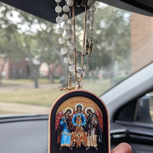 May include: A car hanging ornament featuring a religious icon with angels and a central figure. The icon is framed in a wooden arch shape and is suspended by a beaded chain and a leather cord. The background is blurred.