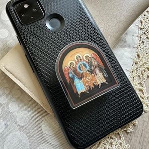 May include: A black smartphone case with a textured geometric pattern on the back, featuring a centered sticker depicting a religious icon of three figures with halos and wings, rendered in traditional colors of red, blue, and gold with dark borders.