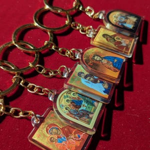 May include: A selection of gold-coloured keyrings, each displaying a religious icon within a clear, rectangular pendant. The icons depict various religious figures in vibrant colours. The keyrings are arranged on a red surface.